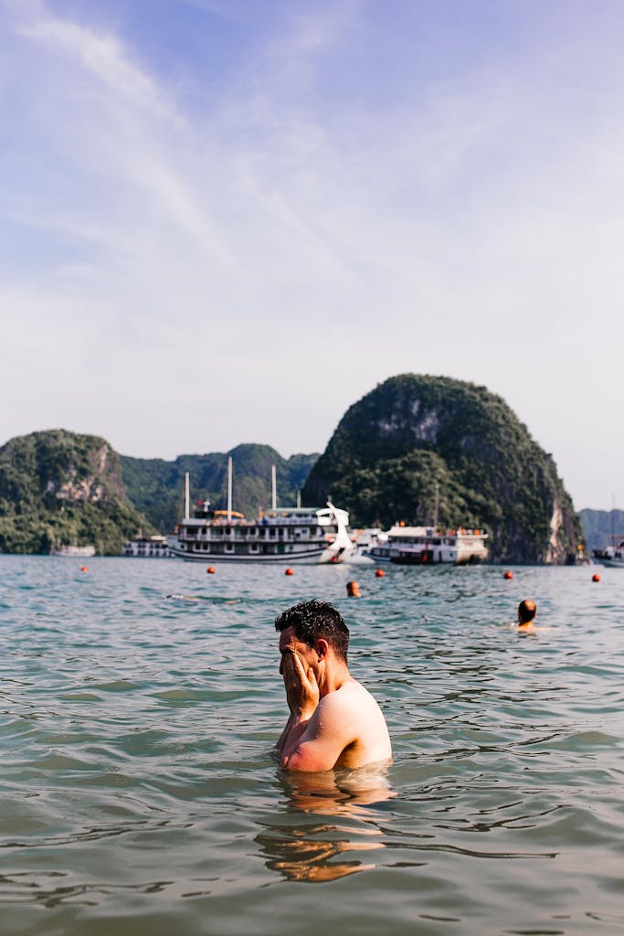 A man enjoys the clear waters of Ha Long Bay, with cruise boats and limestone formations in the background.