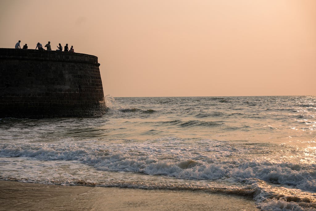 Silhouetted figures on a historic fort wall during a peaceful Goa sunset by the sea.